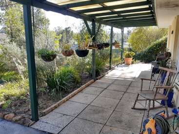 A covered patio with hanging plants overlooks a lush garden. There are wooden chairs, a potted plant, outdoor furniture, and a garden hose neatly coiled nearby.