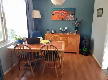 A cozy dining room with a wooden table, four chairs, laptop, potted plant, sideboard, eclectic art, flowers, window, floor lamp, and speaker.
