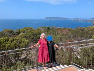 Two women stand on a wooden platform overlooking a vast blue ocean and green coastline. The sky is clear, and a distant island is visible.
