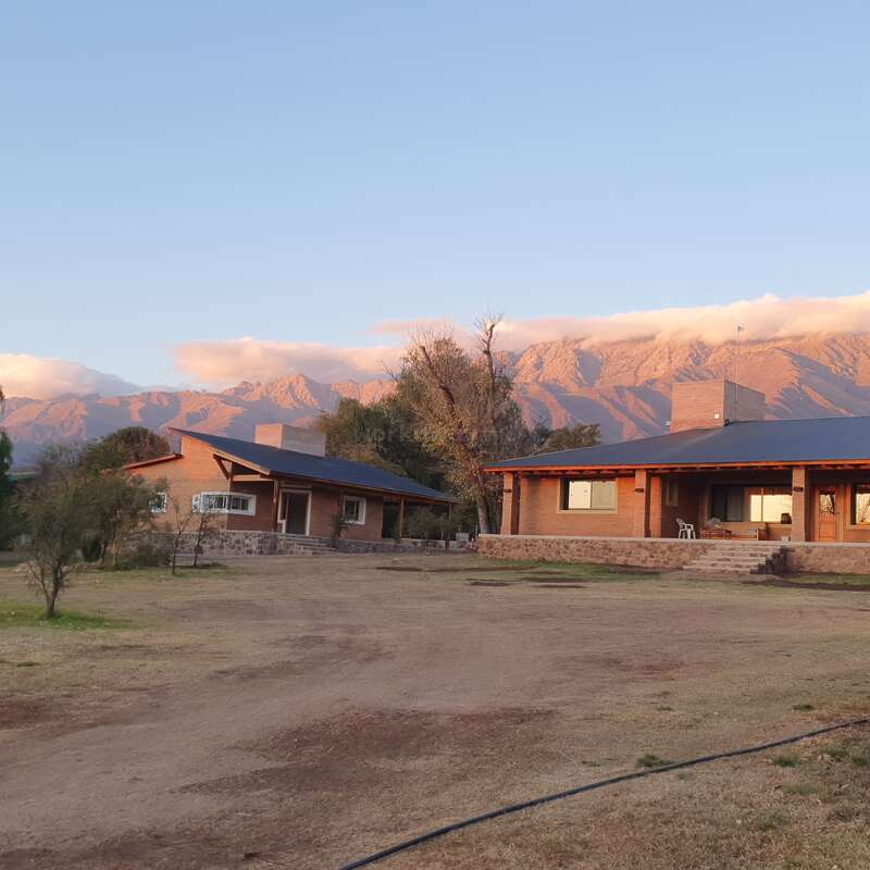The image depicts a single-story house with a flat roof, situated in front of a mountain range, featuring a large yard and a serene atmosphere.