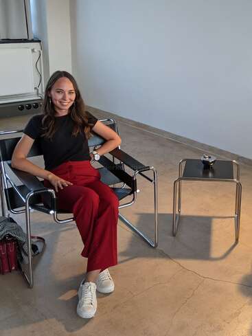 Une femme est confortablement assise sur une chaise moderne ; elle porte un t-shirt noir et un pantalon rouge. Elle sourit, avec une table et un sac à côté d'elle.