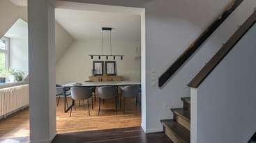 Une salle à manger moderne, avec six chaises grises et une table rectangulaire, est éclairée par un luminaire contemporain. Le parquet et l'escalier apportent une touche chaleureuse. Des plantes près de la fenêtre.