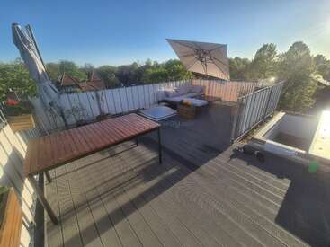 Une terrasse ensoleillée sur le toit, agrémentée de mobilier en bois, d'un grand parasol, d'un coin salon confortable avec des coussins, de plantes en pot et d'arbres verdoyants en arrière-plan. Une atmosphère propice à la détente.
