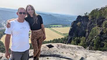 Un homme et une femme posent sur une falaise rocheuse, entourés d'arbres et de champs verdoyants, avec en arrière-plan des collines lointaines et un ciel clair et lumineux.