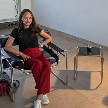Uma mulher está sentada confortavelmente em uma cadeira moderna, vestindo uma blusa preta e calças vermelhas. Ela está sorrindo, com uma mesa e uma bolsa ao seu lado.
