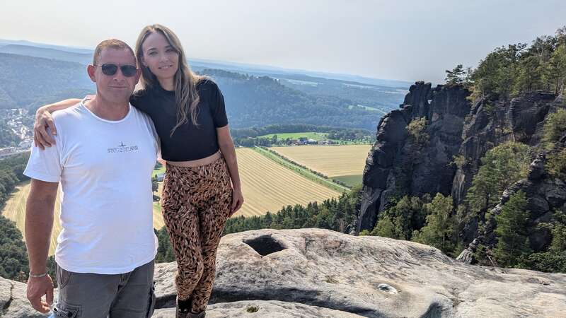 Un homme et une femme posent sur une falaise rocheuse, entourés d'arbres et de champs verdoyants, avec en arrière-plan des collines lointaines et un ciel clair et lumineux.