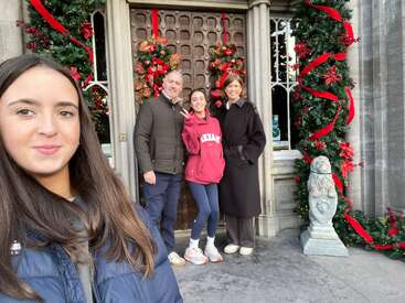 Une famille pose devant une porte joliment décorée de guirlandes de Noël et de rubans rouges. Une petite fille prend un selfie tandis que les autres sourient derrière elle.