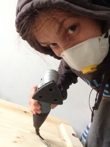 A person wearing a hoodie and a protective mask is holding a power sander, working on a light wooden surface, possibly sanding it for a project.