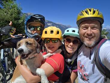 Une famille souriante portant des casques de vélo pose en plein air avec son chien. Journée ensoleillée, montagnes et ciel bleu en arrière-plan. Tout le monde a l'air heureux, prêt pour l'aventure.