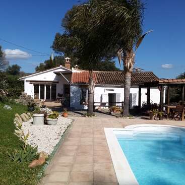 La imagen muestra una serena casa blanca con tejado de tejas marrones, rodeada de una piscina, palmeras y un frondoso jardín, bajo un cielo azul despejado.