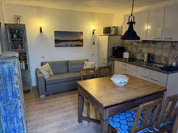 Cet espace de vie confortable comprend un canapé gris, une table à manger en bois avec des chaises, une kitchenette, des étagères rustiques, des décorations murales et un éclairage chaleureux, créant ainsi une atmosphère accueillante.