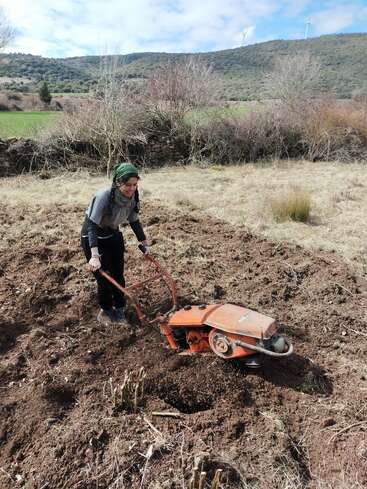 Eine Frau bearbeitet mit einer orangefarbenen Fräse den Boden auf einem Feld. Sie ist warm gekleidet, trägt ein grünes Kopftuch und ist von einer ländlichen Landschaft umgeben.