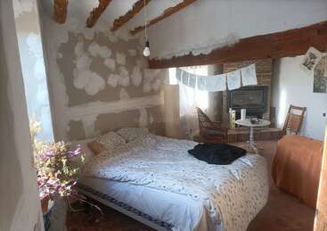 Ein gemütliches, rustikales Schlafzimmer mit freiliegenden Balken, unfertigen Wänden, einem Metallbett, gemusterter Bettwäsche, Wäschetrockner, Holzstühlen, Topfpflanzen, Kamin und einfallendem Sonnenlicht.