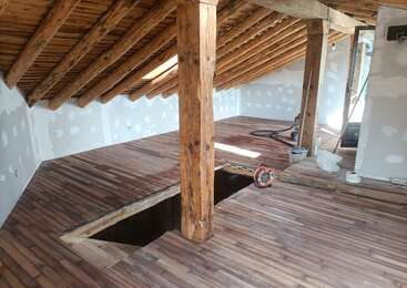 Das Bild zeigt einen Raum mit einem Holzboden und einer Holzdecke, in der ein großes Loch im Boden und ein Holzbalken in der Mitte zu sehen sind.