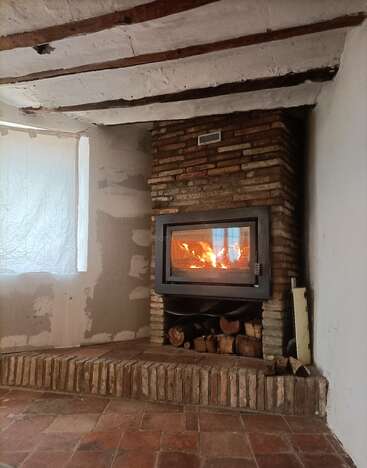 Das Bild zeigt einen rustikalen Raum mit einem Holzofen, einer gemauerten Feuerstelle und einem Fenster mit weißem Vorhang vor einer Kulisse aus freiliegenden Balken und einem Fliesenboden.