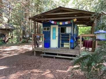 Uma cabana aconchegante e rústica, cercada por árvores altas. Bandeiras coloridas pendem do telhado da varanda. Uma mesa com cadeiras fica do lado de fora, convidando a relaxar na natureza.