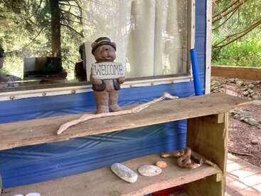Uma pequena estatueta de urso segurando uma placa com a palavra “BEM-VINDO” está em uma prateleira rústica de madeira do lado de fora de um prédio azul, cercada por pedras, galhos e a natureza.