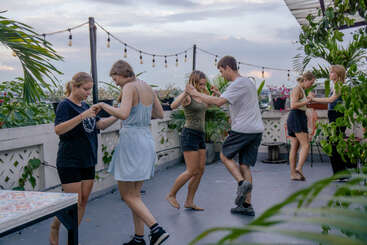 Un grupo de jóvenes baila en una azotea rodeada de plantas, bajo luces de cuerda, disfrutando de un ambiente relajado y social al caer la tarde.