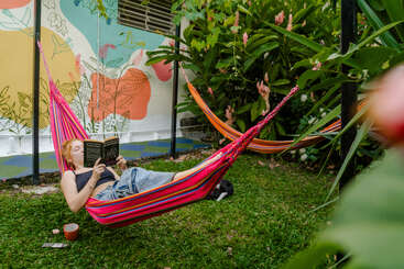 Una mujer se relaja en una hamaca de colores, leyendo un libro en un exuberante jardín con plantas vibrantes, una taza y un colorido mural en la pared.
