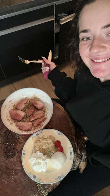 A smiling woman holds a fork and knife, sitting by a wooden table with plates of cooked meat, sauerkraut, tomatoes, and what appears to be cottage cheese.