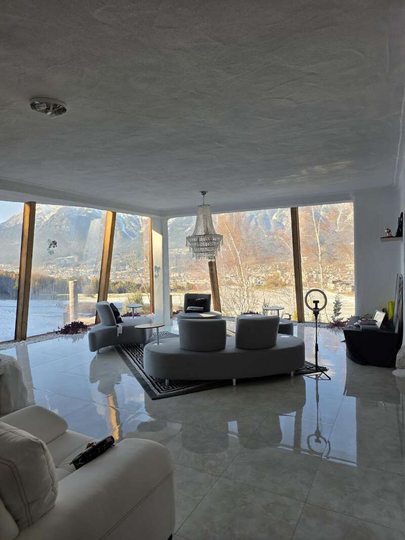 A modern living room with large glass windows showcases a stunning mountain view. Cozy gray and white furniture, chandelier, and luxurious tiles create an elegant, inviting space.