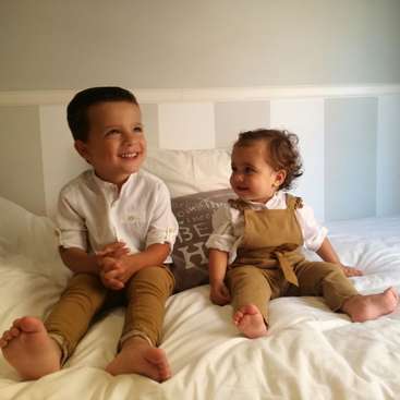 L'image représente deux jeunes enfants, un garçon et une fille, assis sur un lit aux draps blancs, tous deux vêtus d'une chemise blanche et d'un pantalon ou d'une salopette de couleur beige.