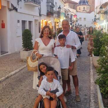 Uma família de cinco pessoas, vestida de branco e bege, posa em uma charmosa rua de paralelepípedos com arquitetura mediterrânea, plantas e iluminação aconchegante, desfrutando de uma noite agradável.