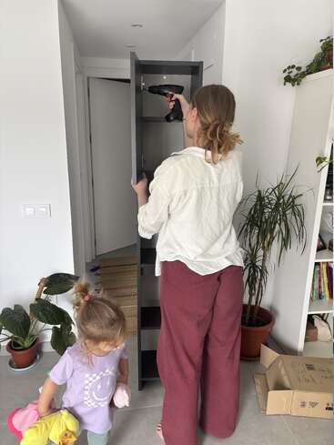 Eine Frau baut mit einer Bohrmaschine ein Regal zusammen, während ein junges Mädchen mit Stofftieren daneben steht. Mehrere Topfpflanzen schmücken den Raum.