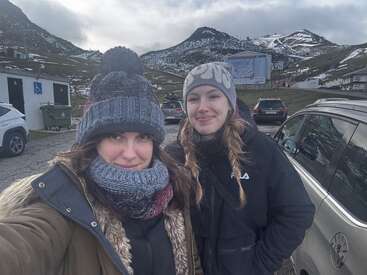 Duas mulheres com chapéus e jaquetas de inverno sorriem para uma selfie em uma área montanhosa com neve, cercadas por carros estacionados e céu nublado ao fundo.