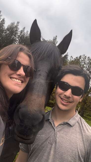 Une femme et un homme portant des lunettes de soleil sourient, avec un cheval noir entre eux. Ils se trouvent en plein air, sous un ciel nuageux, avec des arbres en arrière-plan, et profitent ensemble de ce moment.