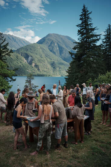 Un grupo de personas se reúne al aire libre cerca de un lago y unas montañas, compartiendo comida en una mesa de picnic, rodeadas de árboles y bajo un cielo azul brillante.