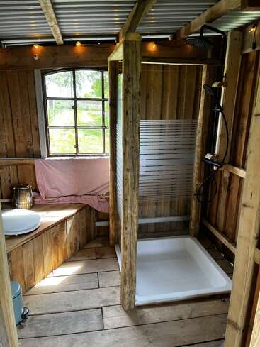 Cette salle de bains rustique présente des murs et un sol en bois, une douche avec une cloison en verre, des guirlandes lumineuses, une fenêtre avec vue sur l'extérieur et un banc confortable avec un tissu rose.