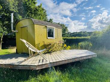 Une petite cabane jaune est posée sur une terrasse en bois dans une prairie verdoyante, avec une chaise longue, une table, des chaises, des arbres et un ciel d'un bleu éclatant.