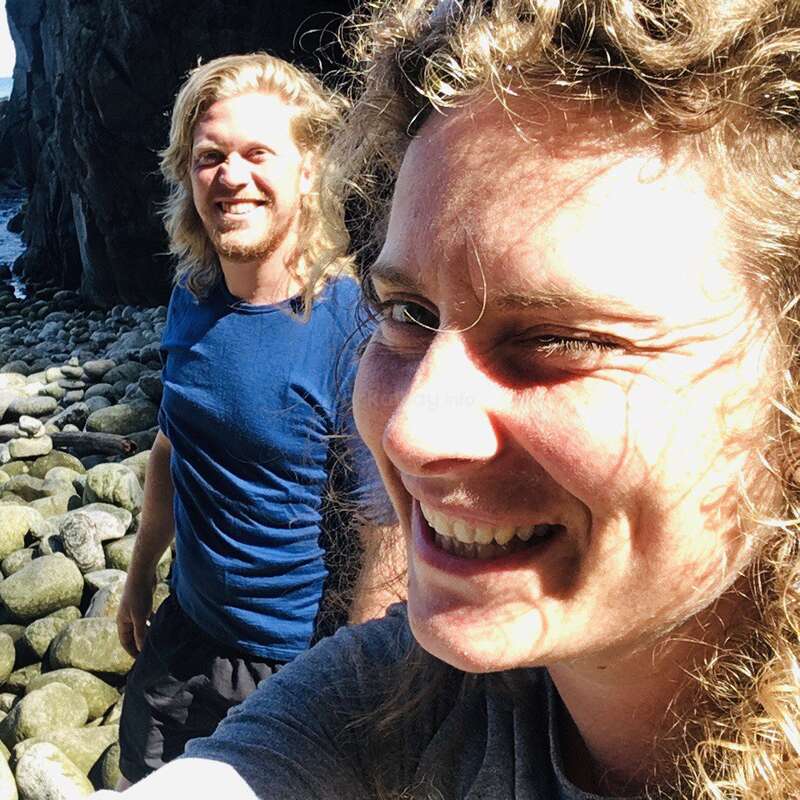 Une femme souriante prend un selfie en gros plan tandis qu'un homme se tient derrière elle. Ils sont en plein air, debout sur un terrain rocailleux près d'une falaise, sous un soleil radieux.