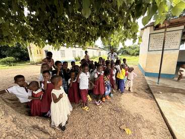 Un groupe de jeunes enfants, pour la plupart en uniforme, se tient sous un grand arbre près d'un bâtiment scolaire, accompagné d'adultes, par une journée ensoleillée.