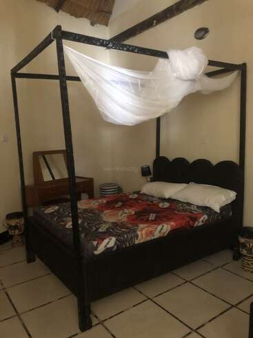 Une chambre confortable avec un lit à baldaquin orné d'une moustiquaire blanche, des draps à motifs floraux, une coiffeuse avec miroir, une décoration de style africain et un sol carrelé.