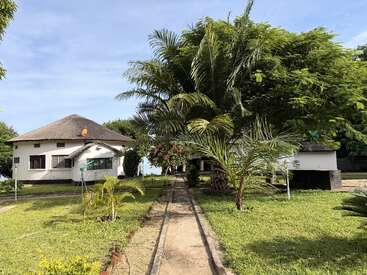 Une scène paisible avec deux bâtiments blancs, des pelouses verdoyantes, des palmiers, un ciel dégagé et un trottoir qui traverse un jardin tropical serein.