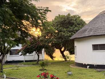 Uma cena tranquila ao pôr do sol, com grama verde, flores coloridas, árvores, prédios brancos e uma cabana com telhado de palha, criando uma atmosfera serena, aconchegante e convidativa ao ar livre.