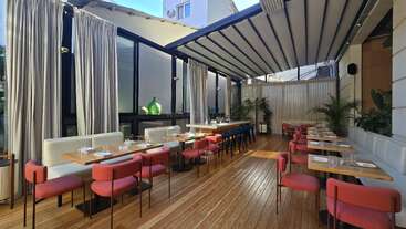 This restaurant features modern decor with wooden floors, red chairs, white cushioned seating, large windows, curtains, plants, elegant tables set, and a partially open roof.