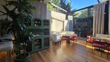 This image shows a modern, sunlit restaurant with wooden floors, a green cabinet, potted plants, pink chairs, and large glass windows overlooking a garden. Cozy atmosphere.