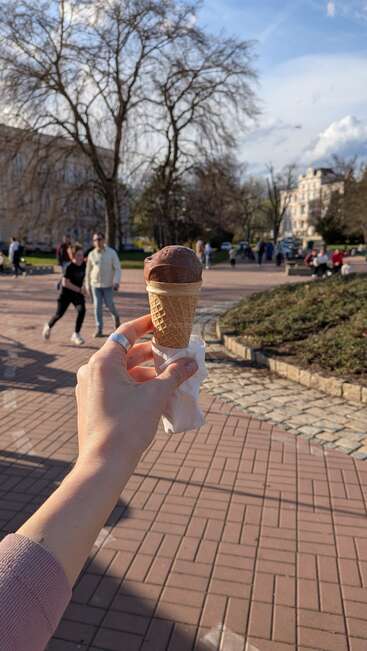 A hand holds a chocolate ice cream cone outdoors on a sunny day. People walk in the background near trees, buildings, and a paved park area.