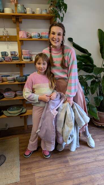 A smiling woman and girl wear matching pink and green striped sweaters, standing in a cozy pottery shop with wooden shelves, plants, and handmade mugs.