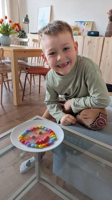 A smiling boy sits beside a glass table displaying a plate of colorful candies arranged in a circle, with melted colors mixing in the center. Cozy home.