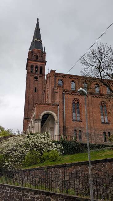 A tall, red-brick church with an elegant spire stands under a gray sky. Blooming shrubs, trees, and a stone fence enhance the atmospheric, slightly moody scene.
