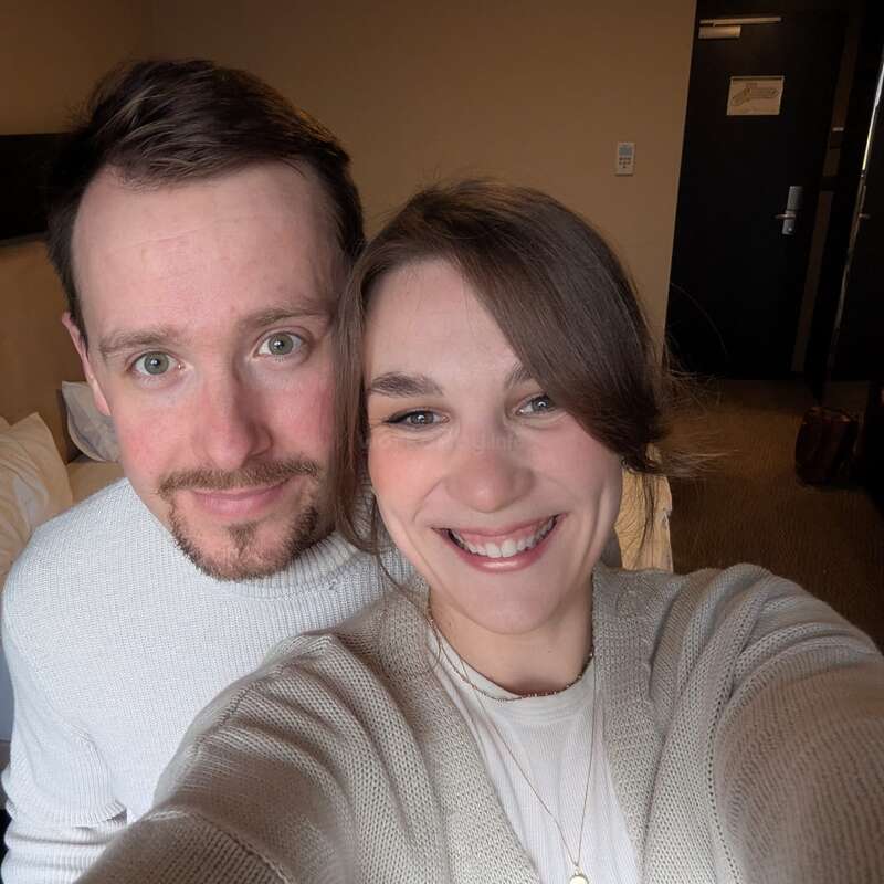 A smiling couple takes a close-up selfie in a cozy, warmly lit room. Both wear light sweaters, radiating happiness and contentment. The background shows a bed and door.
