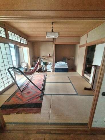 This image shows a traditional Japanese tatami room with sliding shoji doors, a hammock on a rug, a single bed, a shelf, and a fan.