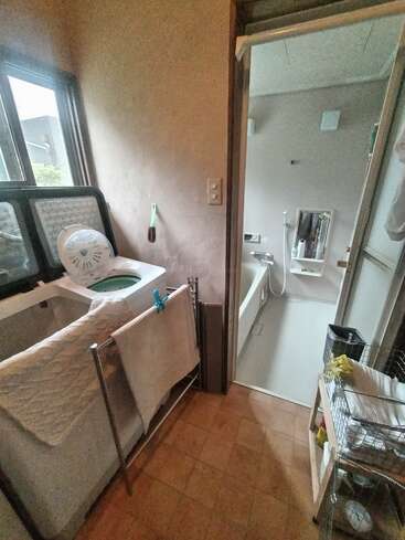 This image shows a small laundry area with a washing machine, drying rack, window, and bathroom entrance with a bathtub, shower, mirror, and organized shelves.