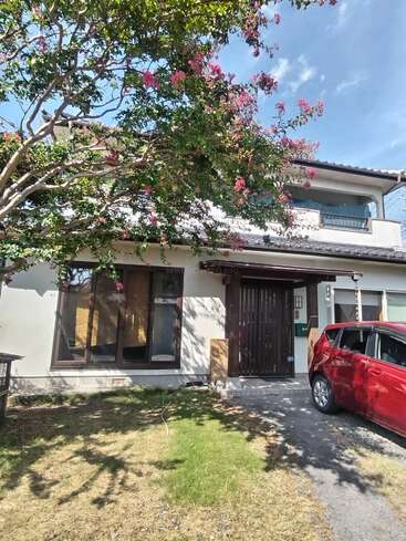 A two-story house with modern and traditional elements, a blooming tree with pink flowers in front, a red car parked on the driveway, sunny day.