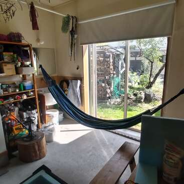 Este acogedor espacio interior cuenta con una hamaca, estanterías con herramientas y suministros, una gran ventana con vistas a un jardín, luz solar que entra a raudales y varios elementos de almacenamiento.