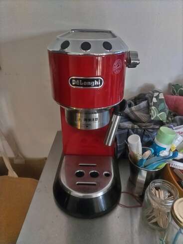 The image shows a red DeLonghi espresso machine on a countertop. Surrounding it are containers holding toothbrushes, toothpaste, and a jar filled with toothpicks.